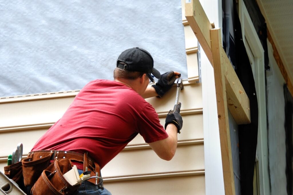 Young roofer installs siding on home. He is holding a hammer and wearing a tool belt.