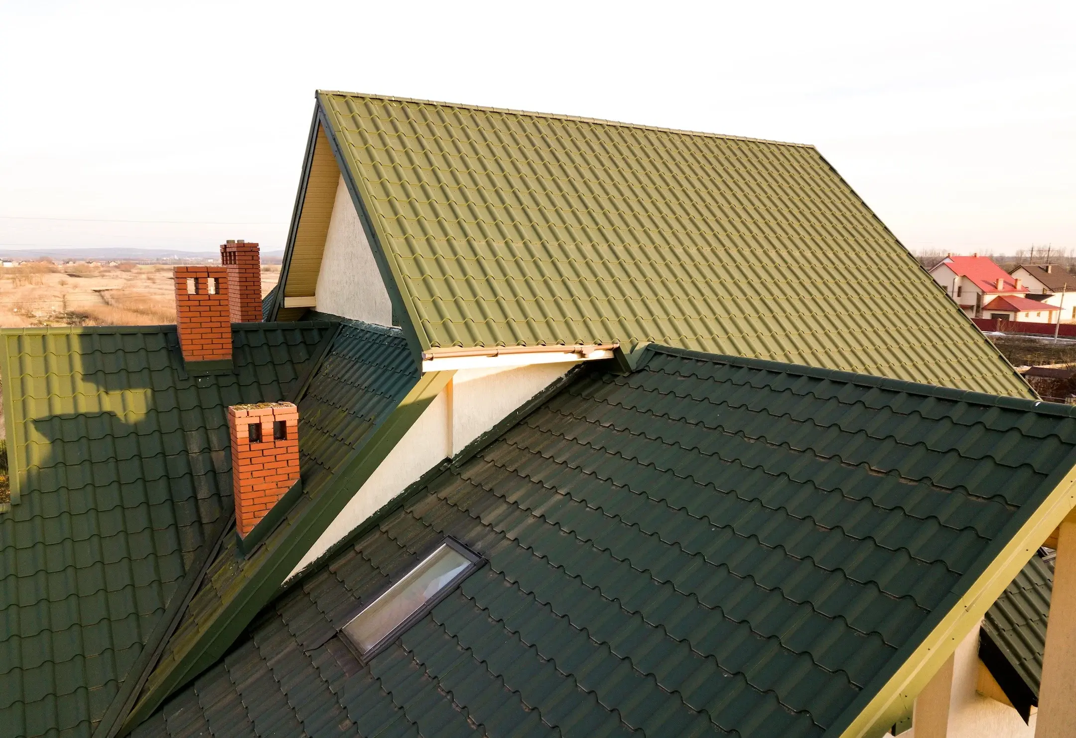 Green metal shingled house roof with attic plastic window and brick chimney.