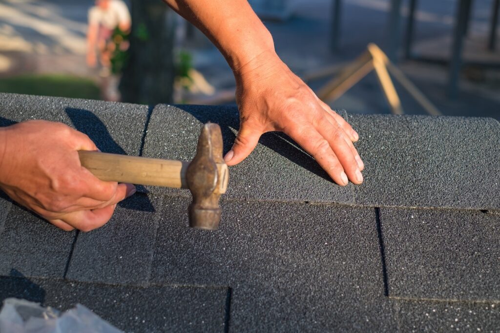 Workers hands installing bitumen roof shingles using hammer in nails.